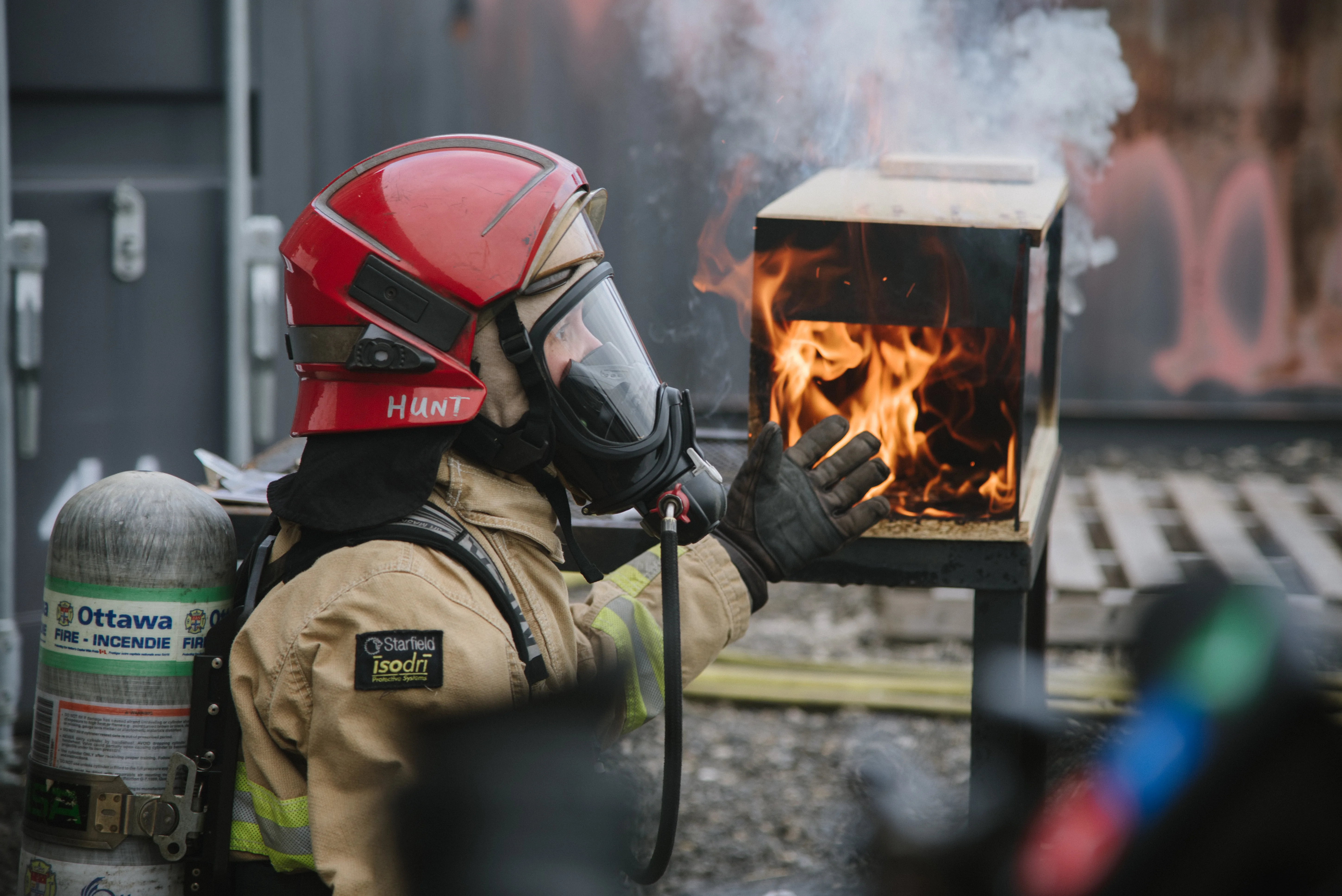An Ottawa Fire Services firefighter in a red helmet observing a small fire-behavior demonstration apparatus